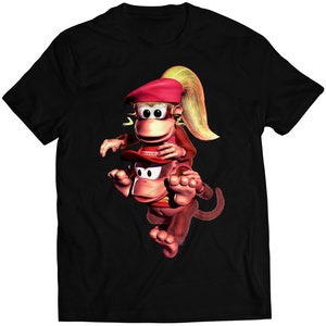 Puede incluir: Camiseta negra con una imagen de dibujos animados de dos monos. El mono de arriba lleva un sombrero rojo y tiene el pelo rubio. El mono de abajo lleva una gorra roja con la palabra "Donkey Kong" en ella.