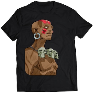 Op de afbeelding: Zwart T-shirt met een cartoonillustratie van een persoon met een bruine huidskleur met rode markeringen op het voorhoofd en een ketting van schedels.