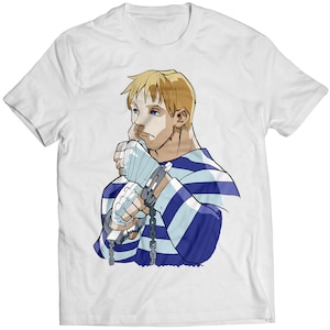 Op de afbeelding: Een wit T-shirt met een cartoonillustratie van een man met blond haar die een blauw-wit gestreept shirt en handboeien draagt. De man kijkt naar beneden en heeft een serieuze uitdrukking op zijn gezicht.
