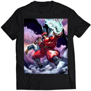 Op de afbeelding: Een zwart T-shirt met een grafische print van een rood gekleed personage met een blauwe energie-explosie, die vecht tegen een ander personage in een witte outfit. Het personage draagt een rode cape en heeft een donkerpaarse achtergrond.