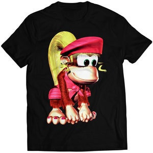 Puede incluir: Camiseta negra con una imagen de dibujos animados de una mona con pelo rubio, que lleva un sombrero rojo y un atuendo rosa y rojo.