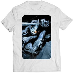 Op de afbeelding: Een wit T-shirt met een grafische print van een blauwe en grijze figuur die een zwaard vasthoudt. De figuur zit in een hurkende positie en de achtergrond is donker.