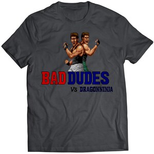 May include: A dark grey t-shirt featuring a pixelated image of two characters from the video game Bad Dudes Vs. DragonNinja. The text "BAD DUDES VS DRAGONNINJA" is printed in red, blue, and white.