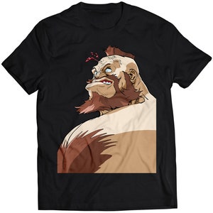 Op de afbeelding: Een zwart T-shirt met een cartoonillustratie van een gespierde man met een baard en een mohawk. De man kijkt omhoog en naar rechts, en zijn uitdrukking is fel.