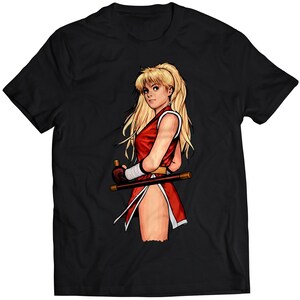 Könnte beinhalten: Ein schwarzes T-Shirt mit einer bunten Illustration einer Frau mit langen blonden Haaren, die ein rot-weißes Outfit trägt und einen Stab hält.