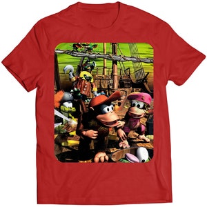 Puede incluir: Una camiseta roja con un estampado gráfico colorido de un grupo de monos de dibujos animados en un barco pirata. Los monos llevan diferentes atuendos y sombreros. El barco navega en un mar verde con un cielo azul de fondo.