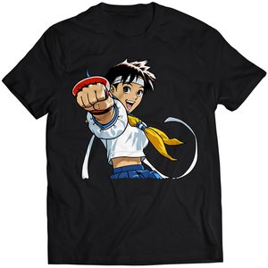 May include: Black t-shirt featuring a cartoon illustration of a female character with brown hair, wearing a white shirt and blue skirt, punching forward with a red and white glove.