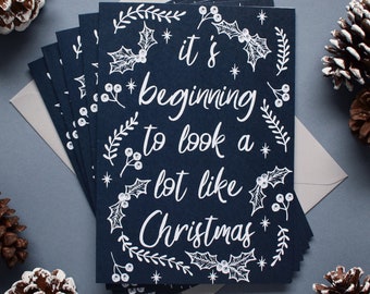 May include: A stack of navy blue Christmas cards with white ink lettering that reads "it's beginning to look a lot like Christmas". The cards are decorated with white holly leaves and berries.