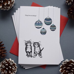 May include: A Christmas card featuring two kittens looking up at a string of blue and white patterned ornaments. The card reads "They're so shiny!"