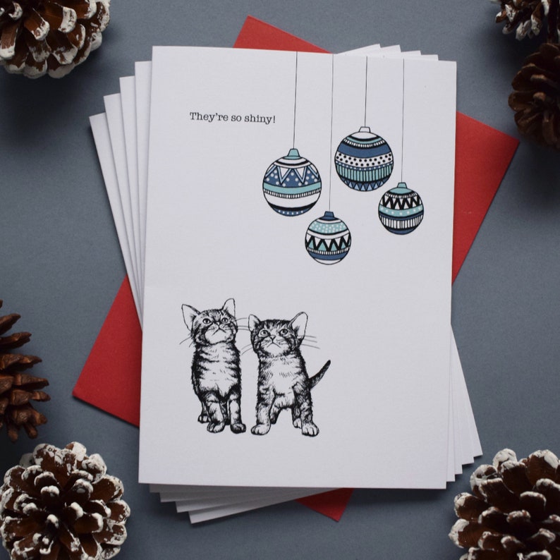 May include: A Christmas card featuring two kittens looking up at a string of blue and white patterned ornaments. The card reads "They're so shiny!"