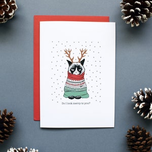 Puede incluir: Una tarjeta de Navidad con un gato gruñón que lleva un suéter verde y rojo con cuernos de reno. El gato está rodeado de un fondo blanco con pequeños puntos grises. El texto en la tarjeta dice "Do I look merry to you?"