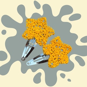 Op de afbeelding: Twee gele gehaakte haarclips in de vorm van een ster. De stervormige clips zijn gemaakt van een getextureerd, open geweven garen en zijn bevestigd aan zilveren metalen klikclips. De achtergrond is lichtbeige met grijze en witte abstracte vormen.