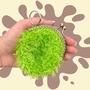 May include: A round, lime green, fluffy coin purse with a metal clasp and a silver key ring. The purse has a textured, fuzzy appearance and a decorative metal frame. The background is a light beige with brown abstract shapes.