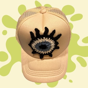 May include: A beige trucker hat featuring a mesh back and a solid front panel. Centered on the front is a circular crochet applique depicting an eye motif with a black pupil, a light blue iris, a white ring, and black radiating lashes.