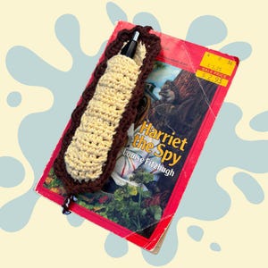 May include: A handmade crochet bookmark in beige and brown, holding a black pen, rests on a vintage copy of "Harriet the Spy" by Louise Fitzhugh. The book has a red cover with a price tag.
