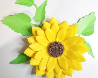 May include: A large paper sunflower with yellow petals and a brown center. The sunflower is surrounded by green paper leaves.
