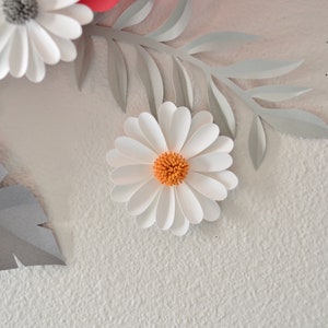 Op de afbeelding: Witte papieren bloemen met grijze papieren bladeren op een witte, textuurrijke achtergrond. De bloemen hebben een gelaagd papierontwerp en het midden van de bloem is oranje.