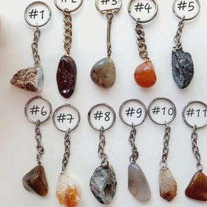 May include: Eleven different keychains with various natural stones attached. The stones are in shades of brown, grey, white, and red. The keychains are numbered 1 through 11.