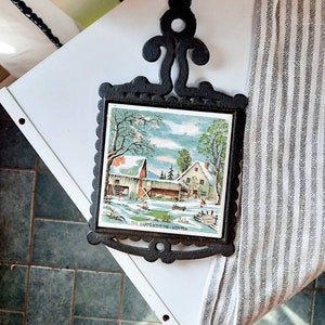 May include: Black cast iron trivet with a white ceramic tile insert featuring a winter scene of a farmhouse with the text "The Farm House - Winter".