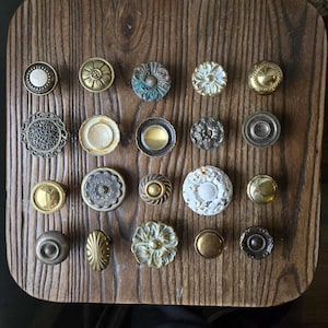 May include: A collection of 18 decorative cabinet knobs in various styles and finishes, including brass, silver, and white. The knobs are arranged on a wooden surface.