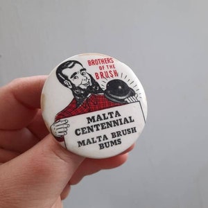 May include: A white button with a red and black design. The button features a cartoon image of a man with a beard holding a sign that reads "Malta Centennial Malta Brush Bums." The text "Brothers of the Brush" is printed above the image.