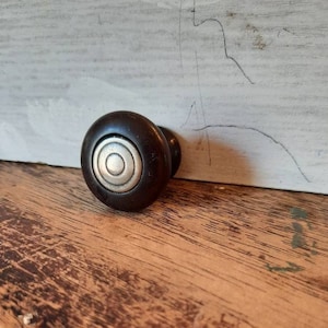 May include: A dark brown cabinet knob with a silver ring around the center. The knob is on a wooden surface.