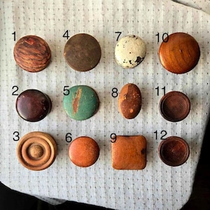 May include: Twelve different knobs in various colors and materials, including wood, metal, and painted finishes. The knobs are arranged in rows on a white fabric surface.