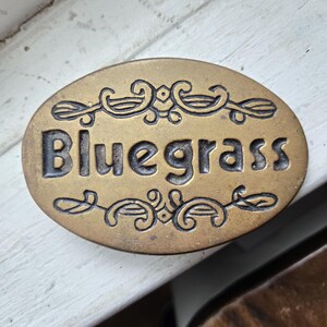 May include: An oval-shaped brass belt buckle featuring the word Bluegrass embossed in the center. The design includes decorative scrollwork patterns above and below the text, with a dark, recessed finish highlighting the lettering and details.