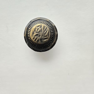 May include: A round, antique-style cabinet knob with a dark bronze finish. The center features a raised, gold-colored floral design within a circular border. The knob has a textured edge and is set against a white background.