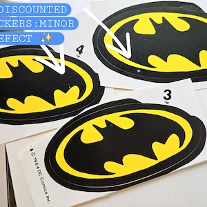 Pode incluir: Tr&ecirc;s autocolantes ovais com o log&oacute;tipo do Batman, apresentando uma silhueta de morcego amarela sobre um fundo preto com uma borda preta. Cada autocolante tem uma pequena aba branca numerada no topo. O texto inclui 1964 DC Comics Inc. e TM.