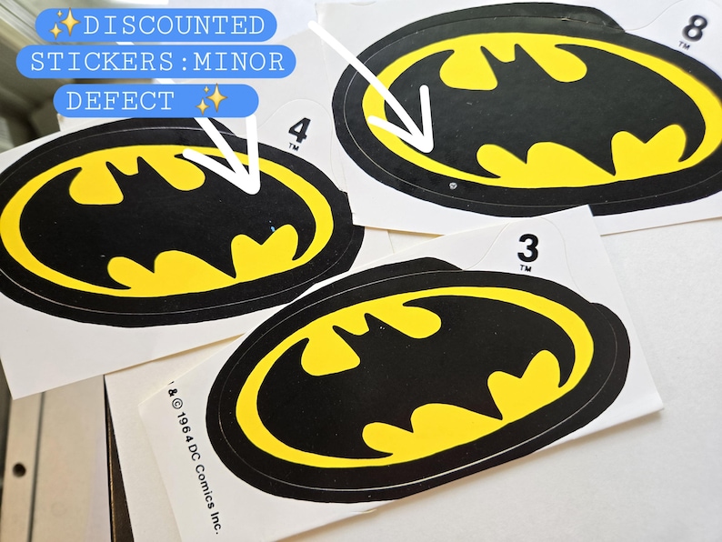 Pode incluir: Tr&ecirc;s autocolantes ovais com o log&oacute;tipo do Batman, apresentando uma silhueta de morcego amarela sobre um fundo preto com uma borda preta. Cada autocolante tem uma pequena aba branca numerada no topo. O texto inclui 1964 DC Comics Inc. e TM.