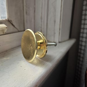 May include: A gold-colored cabinet knob with a round, flat face and a smaller, circular base. The knob has a simple, classic design and is attached to a metal rod. The knob is resting on a white windowsill.