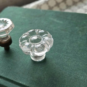 May include: Two clear glass cabinet knobs with distinct shapes. One knob features a rounded, flower-like petal design, while the other is faceted with a starburst pattern on top and a dark metallic base. Both are positioned on a dark green surface.