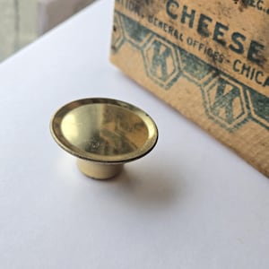 May include: A small, brass-colored knob with a rounded top and a short, cylindrical base. The knob is set against a white background, with a portion of a vintage cheese box visible in the background.