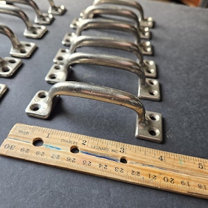 May include: A collection of vintage metal handles with a curved design. Each handle has a rectangular base with two screw holes. A wooden ruler with inch markings is placed next to the handles.