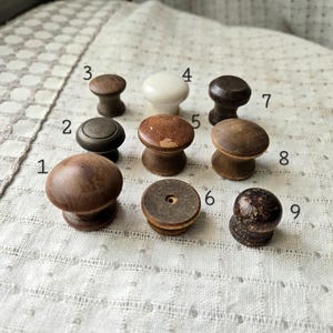 May include: A collection of nine wooden cabinet knobs in various shapes and finishes. The knobs range in color from light to dark brown, with one white knob. The knobs are arranged on a textured, off-white surface.