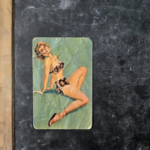 May include: A rectangular vintage-style card featuring a printed illustration of a person with blonde hair wearing a two-piece swimsuit with a dark floral pattern. The person is posed in a seated position against a light green background.