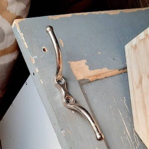 May include: A silver metal hook with two prongs attached to a gray painted wooden surface.