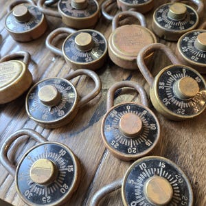 May include: A collection of vintage combination padlocks with brass dials and metal loops. The dials feature black numbers against a white background. The locks are arranged on a wooden surface.