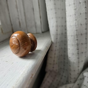 May include: Two wooden cabinet knobs with a warm brown finish rest on a white painted surface. The knobs have a rounded shape with a circular design on the front. A light-colored curtain is in the background.