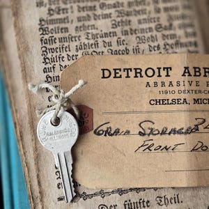 May include: A vintage key from Galesburg, Illinois, attached to a Detroit Abrasive Co. tag with handwritten text. The key and tag rest on the pages of an old book with German text, creating a nostalgic aesthetic.