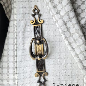 May include: A vertical metal drawer pull with a dark bronze finish and gold-toned accents. The handle features a central textured panel, a swinging loop, and decorative scrollwork at the top and bottom. The text 2-piece is printed in black.