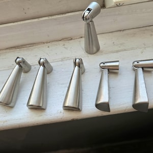 May include: A collection of silver-toned metal cabinet pulls with a unique, angled design. The pulls have a cone-shaped body and a pivoting top section. The hardware is arranged on a white surface, suggesting a focus on home improvement or furniture accessories.