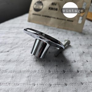 May include: A chrome die-cast knob with a 2-inch diameter, likely for a cabinet or drawer. The knob is shiny and reflective, with a rectangular top and cylindrical base. A vintage paper label is in the background.