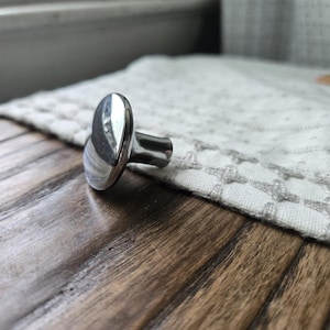 May include: A shiny, silver-colored cabinet knob with an oval head and a short, cylindrical stem. The knob is positioned on a wooden surface, with a textured, off-white fabric in the background.