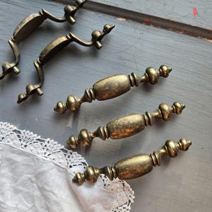 May include: Antique brass cabinet pulls and handles. The handles have a curved design with decorative finials. The pulls have a more elongated shape with a central oval and decorative ends. The hardware is aged with a warm, golden hue.