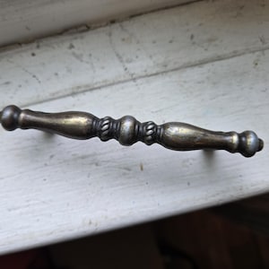 May include: Antique brass-toned drawer pull with a decorative design. The handle features a central section with rope-like detailing and rounded ends. The hardware is set against a white, weathered surface.