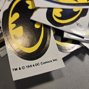 Pode incluir: Uma cole&ccedil;&atilde;o de autocolantes com o log&oacute;tipo do Batman, apresentando uma silhueta de morcego preta centrada num fundo oval amarelo. Cada autocolante inclui o texto impresso TM & &copy; 1964 DC Comics Inc. na margem inferior. Est&atilde;o sobrepostos.