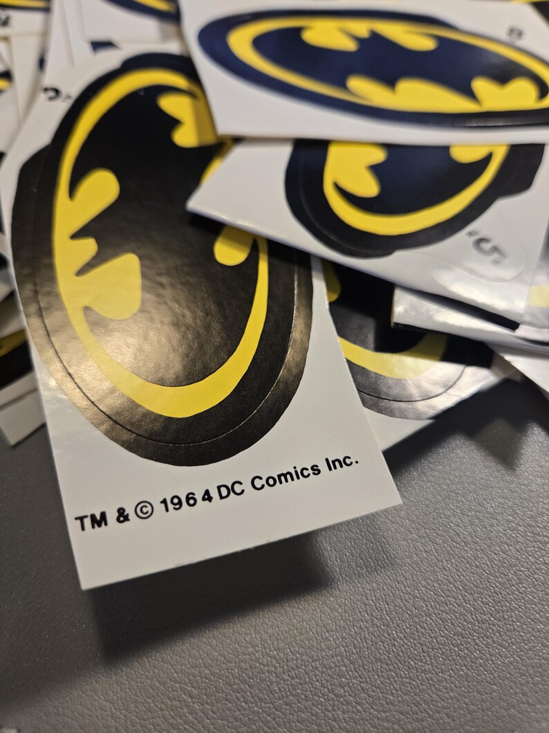 Pode incluir: Uma cole&ccedil;&atilde;o de autocolantes com o log&oacute;tipo do Batman, apresentando uma silhueta de morcego preta centrada num fundo oval amarelo. Cada autocolante inclui o texto impresso TM & &copy; 1964 DC Comics Inc. na margem inferior. Est&atilde;o sobrepostos.