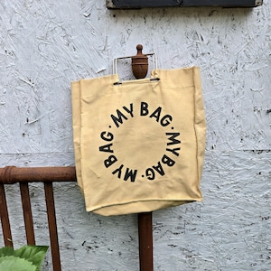 May include: Tan canvas tote bag with the words "MY BAG" printed in a circular pattern in black. The bag has a metal handle and is hanging on a rusty metal rod. The background is a weathered, light gray wooden surface.